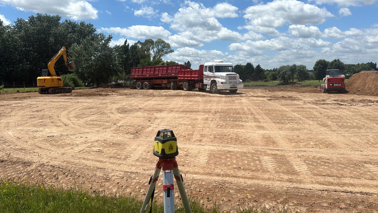 Nivel láser sobre trípode midiendo un terreno nivelado con excavadora y camión al fondo
